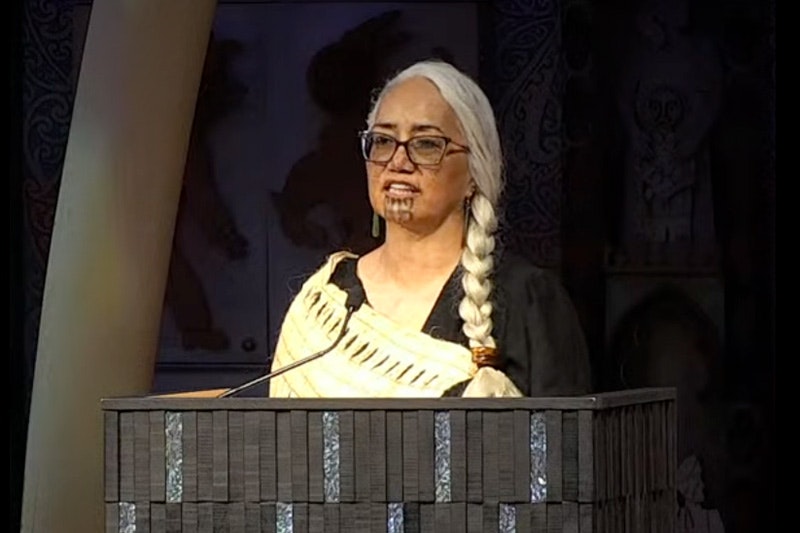 A woman wearing a woven cloak is talking from a podium.