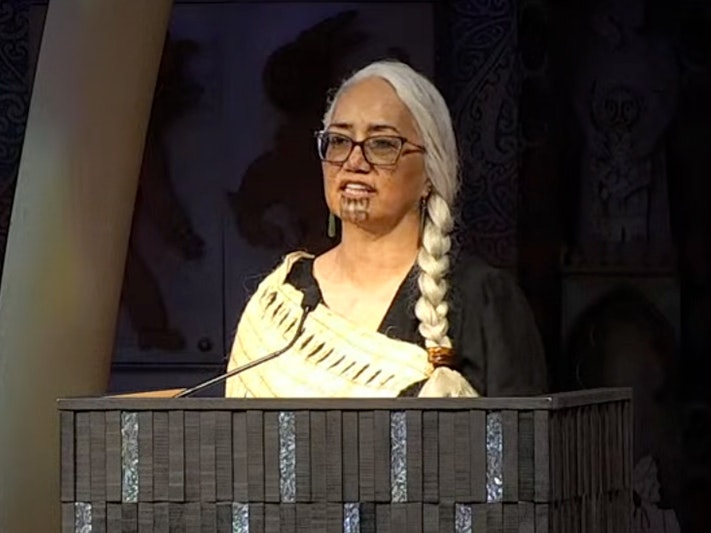 Dr Awhina Tamarapra giving her Volkerling Lecture. 2025 A woman wearing a woven cloak is talking from a podium.