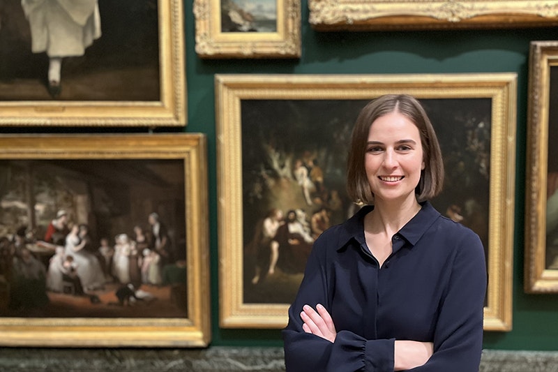 A woman is standing with her arms crossed in front of paintings hung in a gallery.