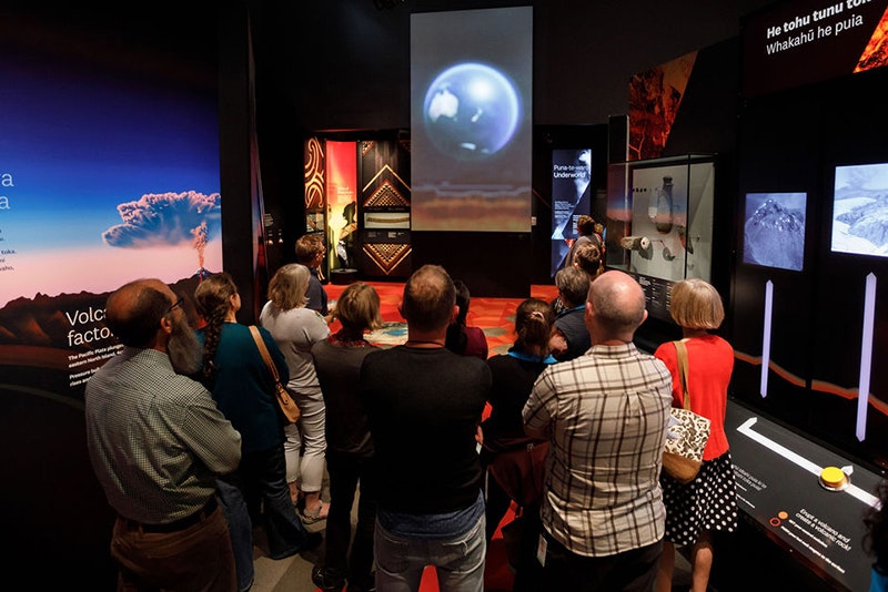Several people standing in a museum space looking at a screen with an image of the world on it, with NZ showing.