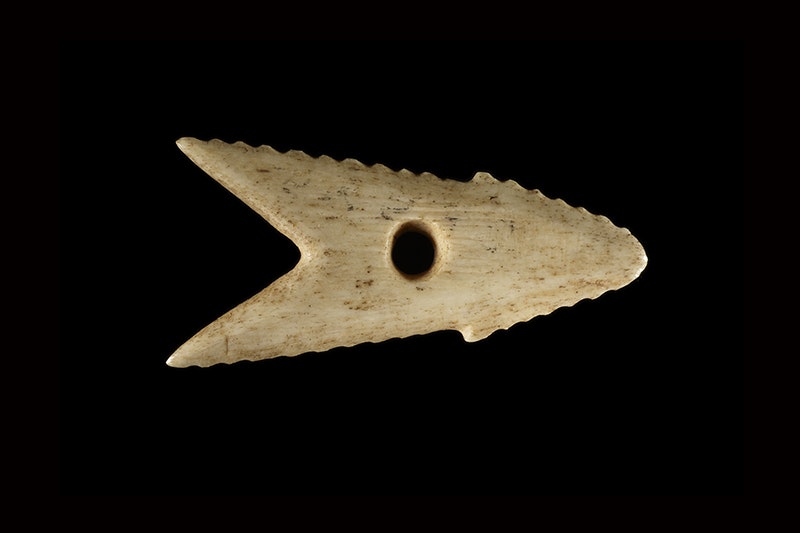 A carved bone harpoon. head in the shape of an arrow pointing right on a black background.