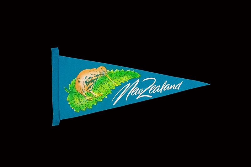 A blue triangular pennant felt flag with the words New Zealand on it, and a kiwi on top of a fern.
