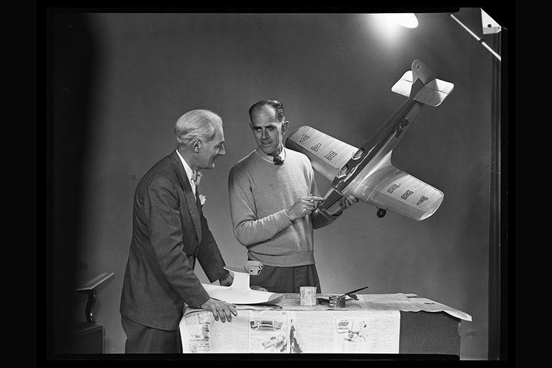 Black and white photo of two men standing at a table with a large light shining on them. One man is holding a model airplane.
