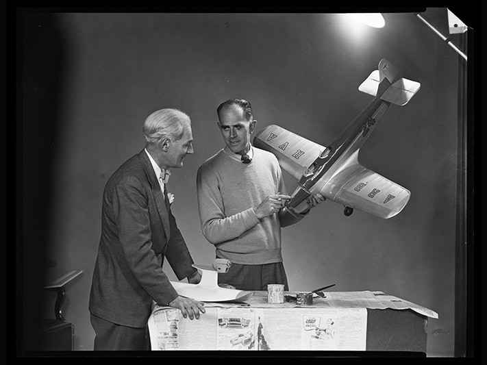 Spencer Digby and R. Bothamly with model plane; 8 August 1952; Spencer Digby Studios. Spencer Digby / Ronald D Woolf Collection. Gift of Ronald Woolf, 1975.Te Papa (C.025860) Black and white photo of two men standing at a table with a large light shining on them. One man is holding a model airplane.