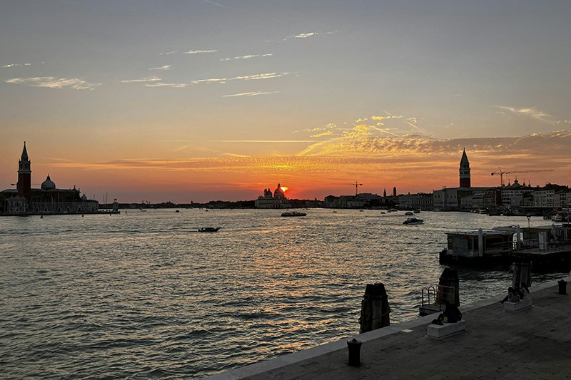 A waterway in Venice with a sunrise or sunset in the background and two of towers on either side of the image.