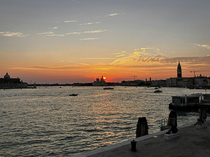 Venice. Photo courtesy of NZ at Venice A waterway in Venice with a sunrise or sunset in the background and two of towers on either side of the image.