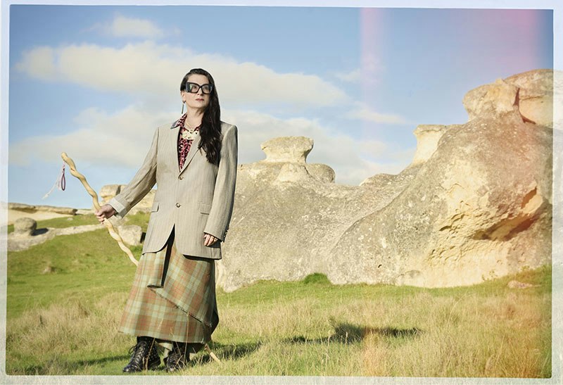 A woman is standing on the side of a hill with rocky outcrops behind her and she is holding a carved walking stick.