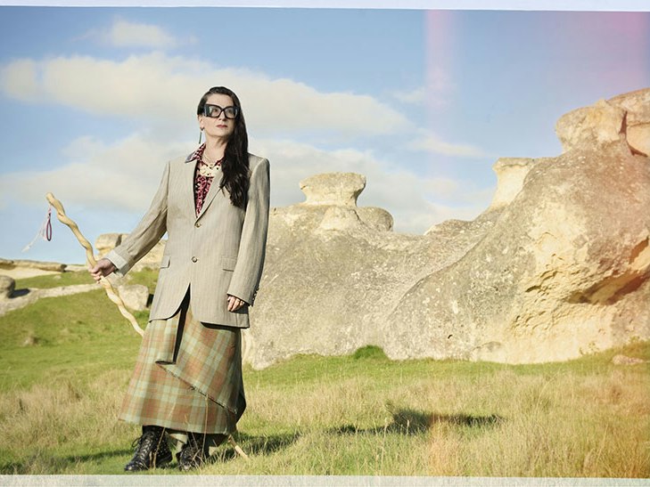 Fiona Pardington. Photo by Meek Zuiderwyk. Courtesy of Creative New Zealand A woman is standing on the side of a hill with rocky outcrops behind her and she is holding a carved walking stick.