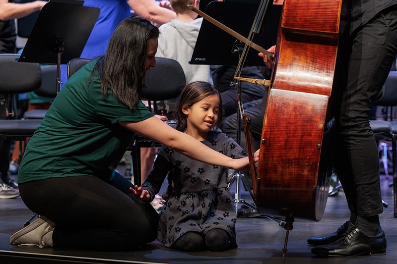 A woman is kneeling on the ground showing a child a cello.