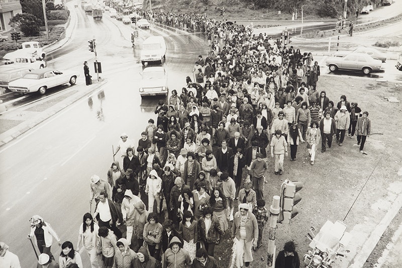 A black and white photo of a lot of people marching along a wet road. There are cars alongside them.