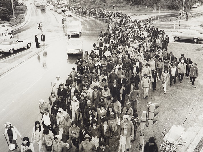 Maoritanga - Scenes from Maori Life. Demonstrations - Maori Land March., Ans Westra, 1975; Wellington. Purchased 1993 with New Zealand Lottery Grants Board funds. Te Papa (O.043304) A black and white photo of a lot of people marching along a wet road. There are cars alongside them.