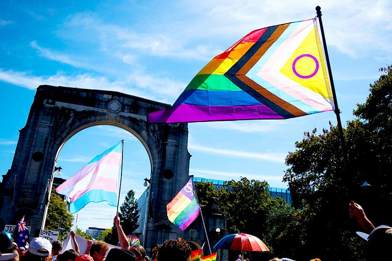 Crowd walks past the Bridge of Remembrance and  Memorial Arch in Christchurch on a sunny day, only the tops of their heads and hats are in frame. Upraised arms wave the Progressive Pride flag, the transgender pride flag, the lesbian pride flag, the New Zealand flag and a rainbow striped umbrella. The sun shines through the flags.