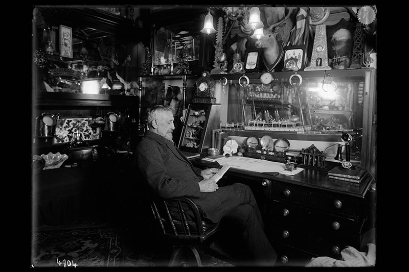 A black and white historical photo of a man in a very crowded office reading something in his hands.