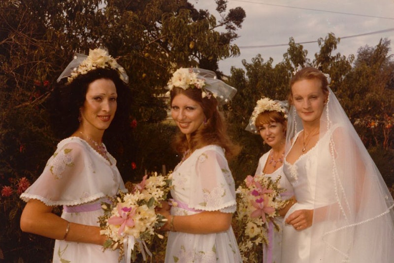 Mid shot of 4 women outdoors. Vanessa wears an elegant white wedding dress with a sculpted sweetheart neckline, a gold locket and long transparent veil. On the left, the 3 bridesmaids are wearing white floral headpieces, empire line dresses patterned with flowers and a lilac waist sash, each holding bouquets. Each bouquet is white with a pink lily in the center.