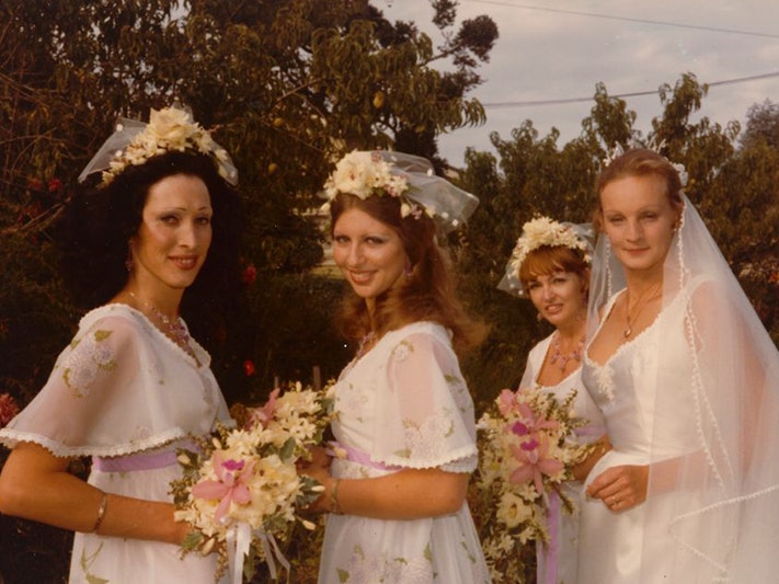 Vanessa Wedding with her bridesmaids, 19 February 1977, Auckland, maker unknown. Gift of Biancha Hartley, 2024. © The copyright holder. Te Papa (O.052287) Mid shot of 4 women outdoors. Vanessa wears an elegant white wedding dress with a sculpted sweetheart neckline, a gold locket and long transparent veil. On the left, the 3 bridesmaids are wearing white floral headpieces, empire line dresses patterned with flowers and a lilac waist sash, each holding bouquets. Each bouquet is white with a pink lily in the center.
