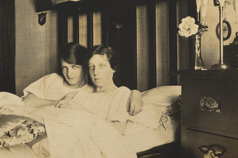 Sepia toned photograph of 2 dark haired women in bed together, looking directly at the viewer. Both wear night gowns and cuddle under a quilted duvet. Kath Whelan (left) has a side swept fringe and a slight smile. Daisy Tinny (left) has a neutral expression. Kath's arms are around Daisy Tinney's shoulders. A flower in a glass vase sits atop a bedside dresser.