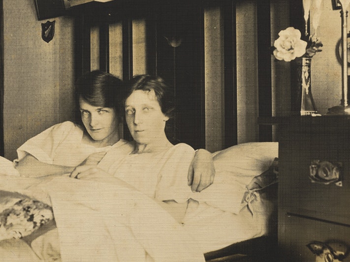 Kath Whelan and Daisy Tinney - self-portrait, 1920s, Wellington, by Daisy Tinney. Te Papa (O.052070) Sepia toned photograph of 2 dark haired women in bed together, looking directly at the viewer. Both wear night gowns and cuddle under a quilted duvet. Kath Whelan (left) has a side swept fringe and a slight smile. Daisy Tinny (left) has a neutral expression. Kath's arms are around Daisy Tinney's shoulders. A flower in a glass vase sits atop a bedside dresser.