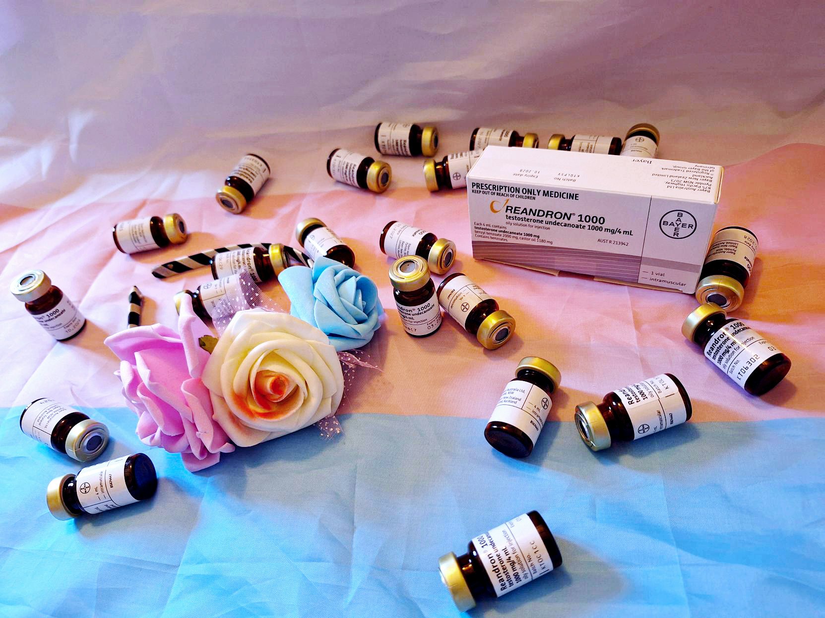 22 small brown, gold capped vials with white labels are scattered across a white, pink and blue striped transgender flag. A headband with foam pink, white and blue roses lies next to the white, rectangular box of Reandron, a common testosterone hormone replacement therapy.