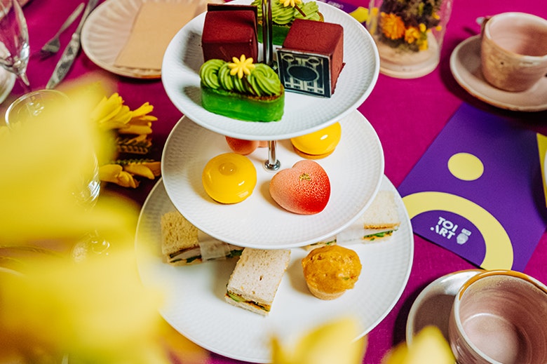 A set of three plates on a stand with different food on each plate, surrounded by a colourful tablecloth and cups and saucers.