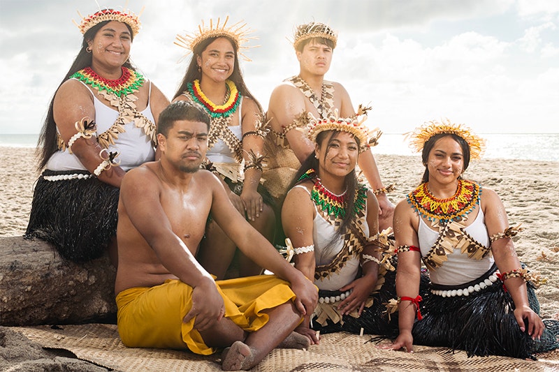 Six people in Kiribati ceremonial clothing sitting on the beach and smiling or looking at the camera.