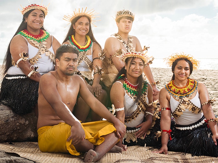 Poster image from Ministry for Pacific Peoples Six people in Kiribati ceremonial clothing sitting on the beach and smiling or looking at the camera.
