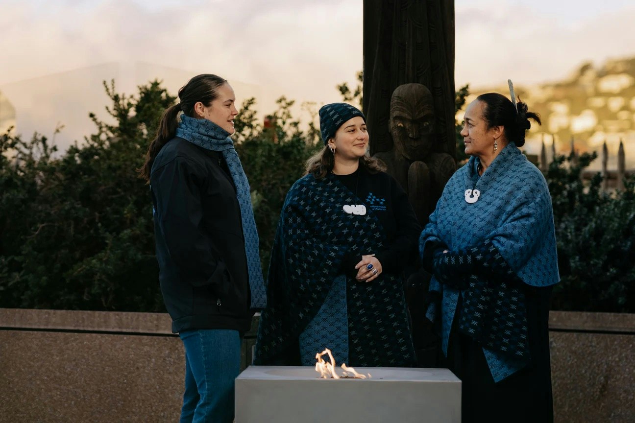 Three women in clothing with the same design are standing next to a fire on a balcony.