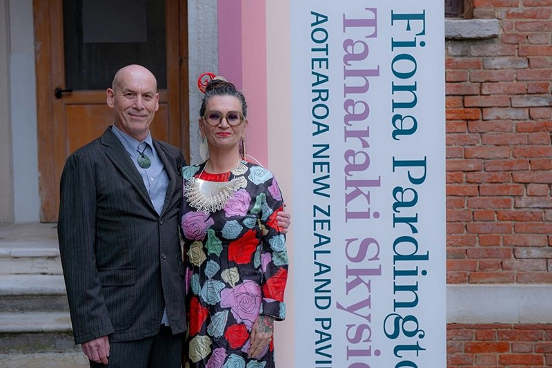Two people standing outside a brick building and looking at the camera.