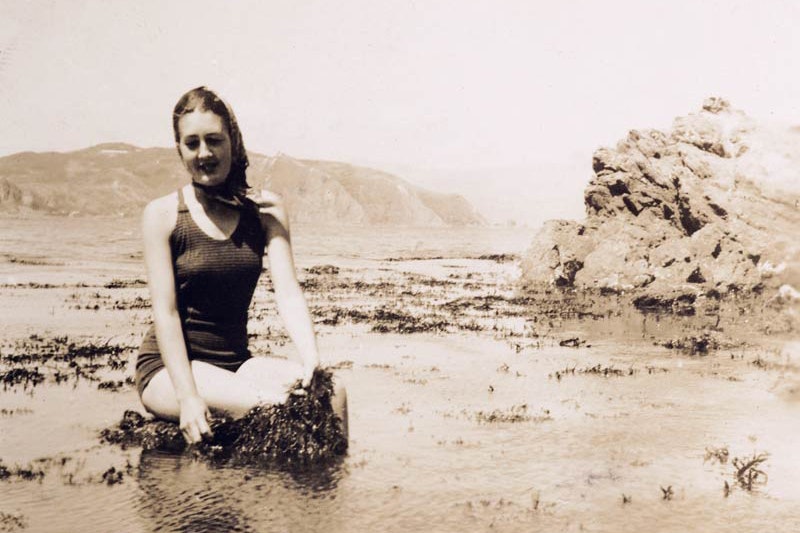 A sepia image of a woman in a bathing suit sitting next to some seaweed on a beach.