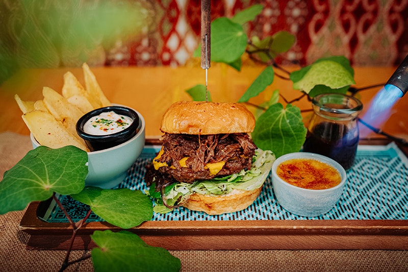 A burger, frieds, a creme brulee dessert and some green leaves are displayed on a wooden tray.
