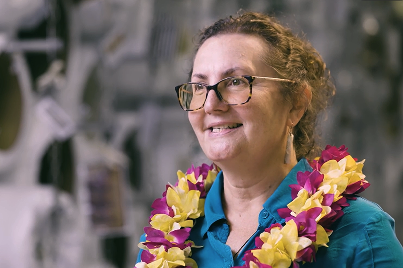 A head and shoulders image of a woman wearing a flower necklace or lei smiling at someone off camera.