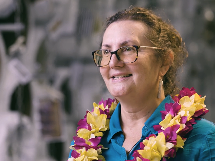 Still from Pauline Reynolds interview. Te Papa A head and shoulders image of a woman wearing a flower necklace or lei smiling at someone off camera.