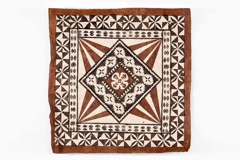 A square tapa cloth with brown and black geometrical patterns painted on it.