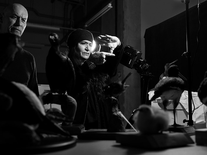 Fiona Pardington and Neil Pardington working in Te Papa's bird store for Taharaki Skyside, by Maarten Holl Te Papa (272962) Two people are in a dark museum store. The woman is framing up a taxidermied bird to take a photograph, the man is looking on.