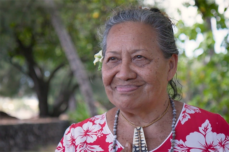 Head and shoulders of a woman with a floral dress and a flower behind her right ear smiling at someone off camera.