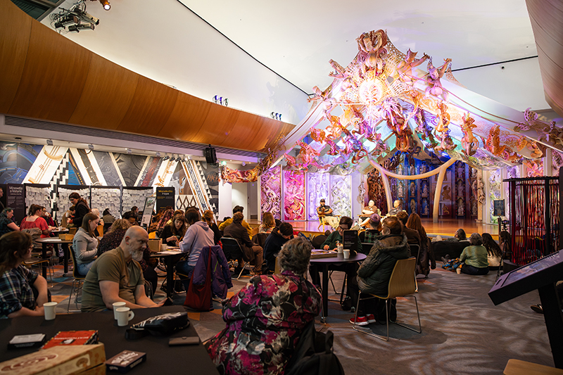 People sitting at tables and on the floor in front of three seated speakers on the raised platform in front of the wharenui at Te Papa.