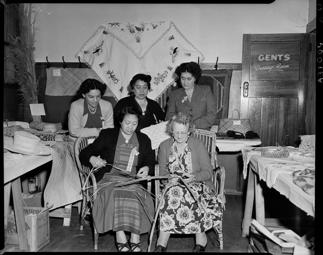 Two women are sitting side by side weaving flax, while three other women look on.