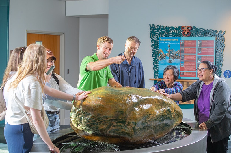 A man in a bright green shirt is sprinkling small stones on a large piece of pounamu that has water spraying on it. He is surrounded by six other people looking on with some of them touching the stone.