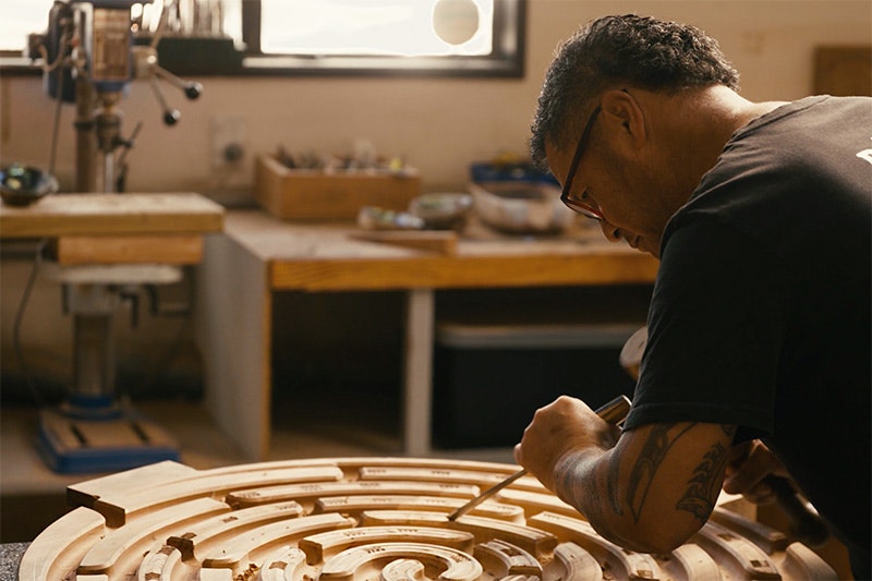 A man is carving a circular disc piece of wood with a chisel and mallet.