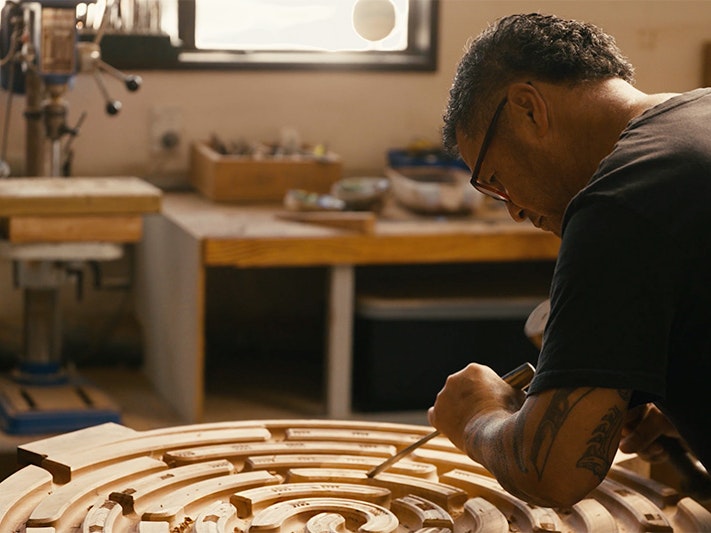 Still from Sam Hauwaho carving Whaka-iro video. Te Papa A man is carving a circular disc piece of wood with a chisel and mallet.