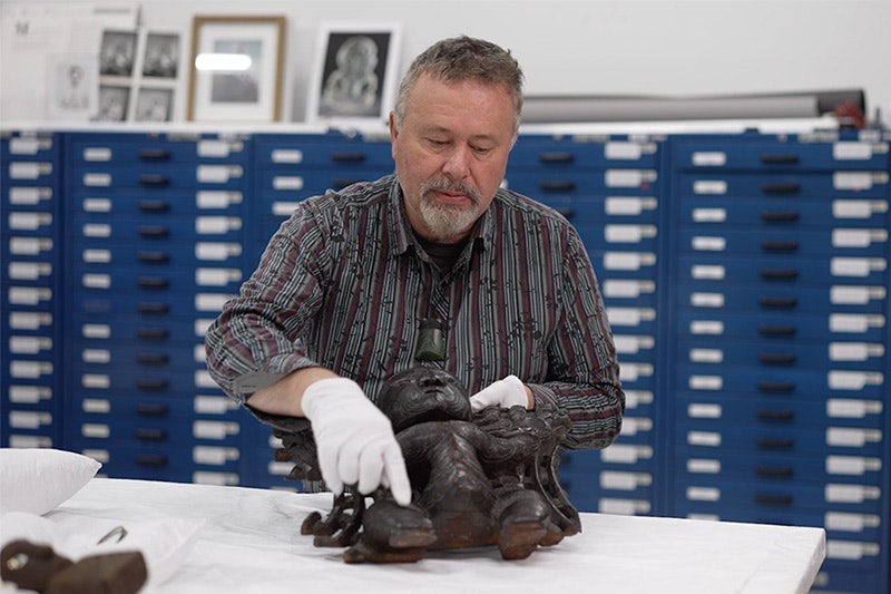 A man is in a museum store room wearing white gloves and holding a wooden carving.
