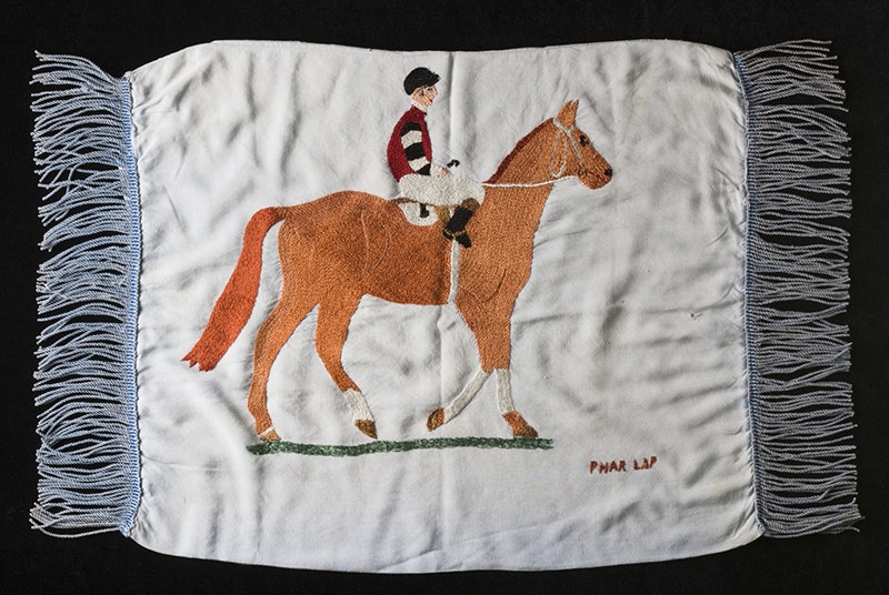 A piece of embroidery with a jockey sitting on a horse and the words PHAR LAP stitched on the bottom-right corner.