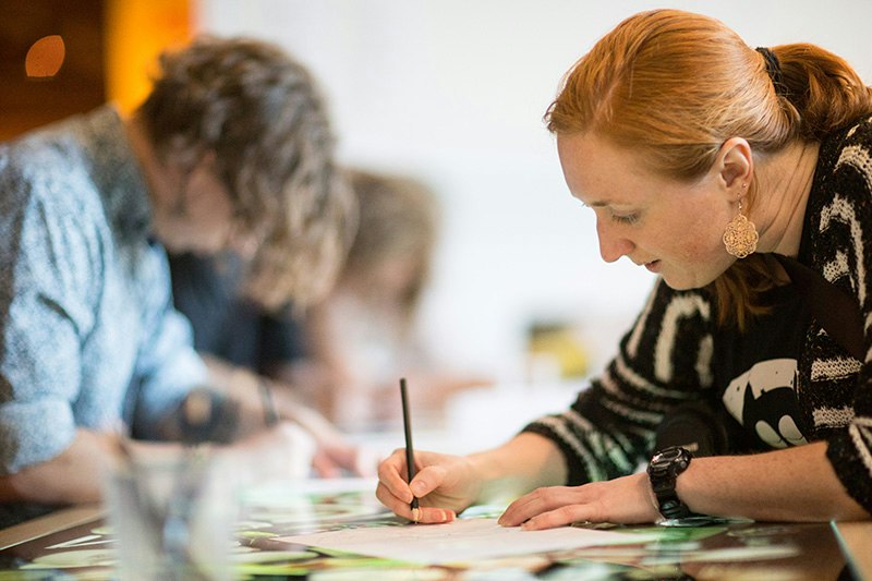 People are working with pencils and paper on a table. There is one woman in focus at the front and other people blurred in the background.