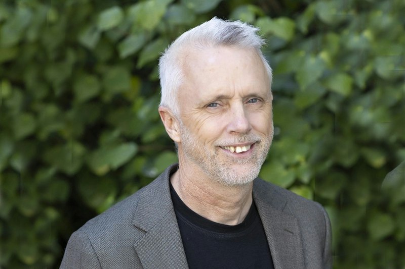 Head and shoulders of a man smiling at the camera. He is standing outside in front of a kawakawa hedge.