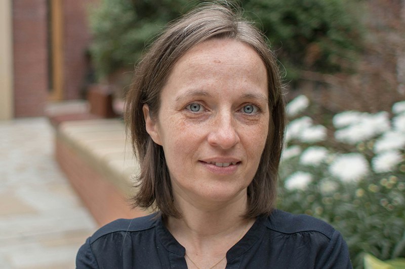 A head and shoulders photo of a woman with shoulder-length hair and blue eyes. She is standing in a brick courtyard.