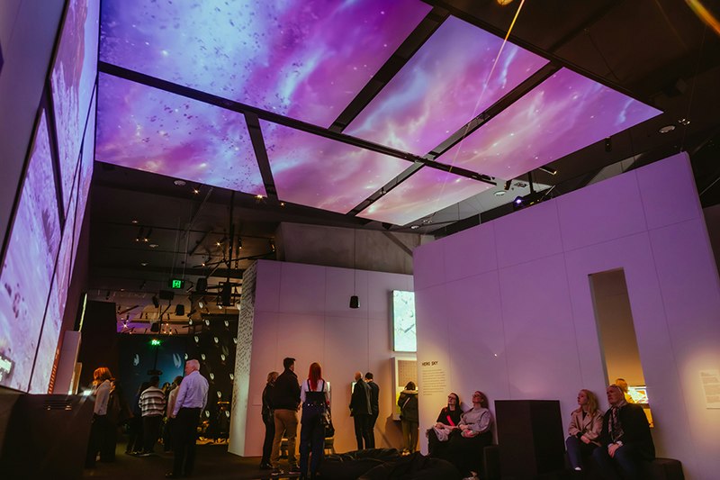 A lot of people in a large room that has a video up one wall and across the ceiling.