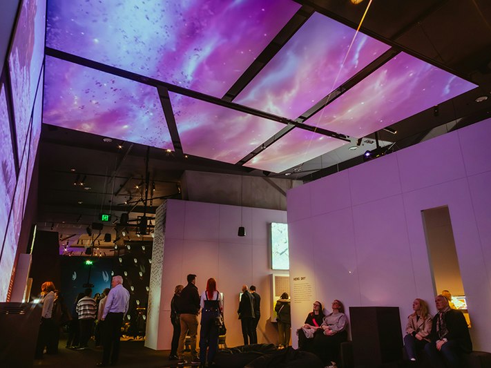 Game Worlds, 2025, ACMI. Photo © Matto Lucas. A lot of people in a large room that has a video up one wall and across the ceiling.