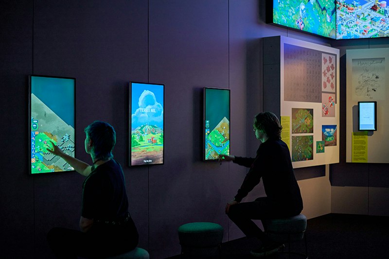Two people are in a darkened gallery. Both are pressing on digital screens mounted on the wall in front of them.