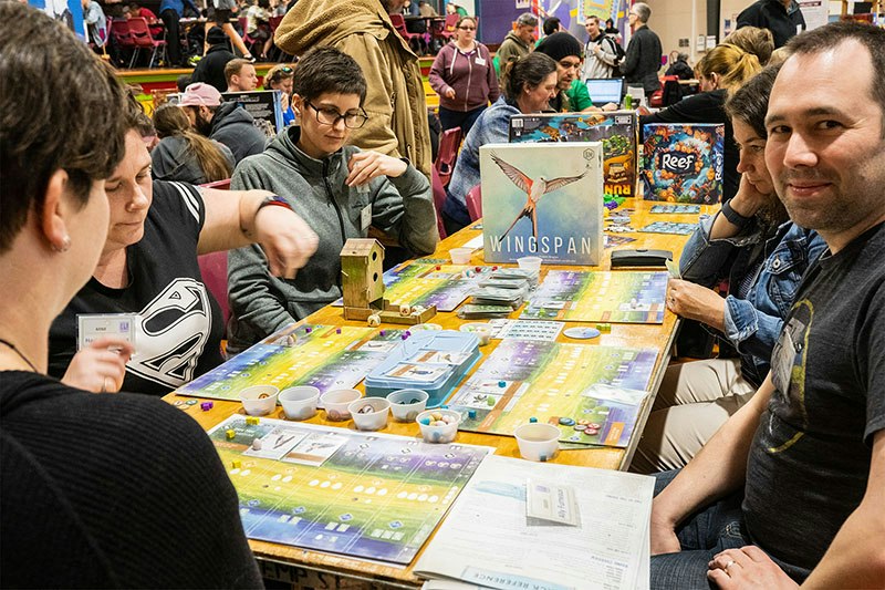A group of people sit in a crowded room around a table playing the board game Wingspan